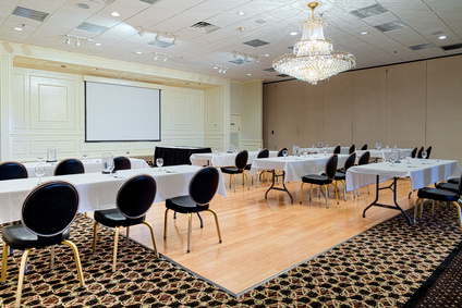 Empty hotel conference meeting or event room provides space for business meetings, conferences, speakers, or events. Tables and chairs set up to veiw projection screen.